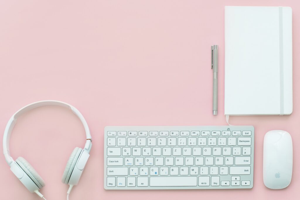 On a pink background is displayed: white headphones, a white keyboard, a white mouse, a white notebook and a grey pen.