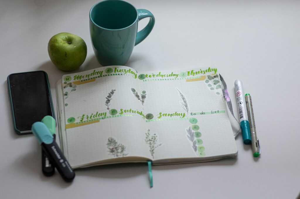 A calendar, phone, pens, apple and cup on a table.